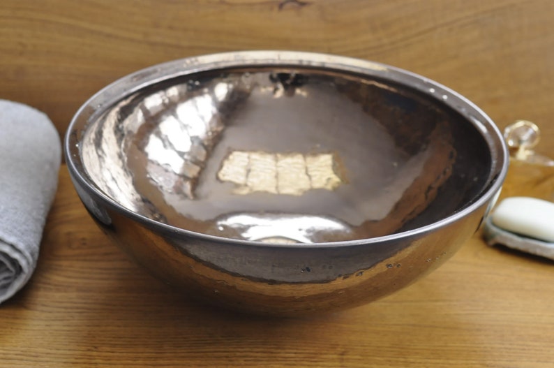 K&ouml;nnte beinhalten: Eine geh&auml;mmerten Metallsch&uuml;ssel mit einem reflektierenden, silberfarbenen Innenraum. Die Sch&uuml;ssel hat eine abgerundete Form und steht auf einer Holzoberfl&auml;che. Ein zusammengerolltes graues Handtuch ist links zu sehen, was auf ein Badezimmer hindeutet.