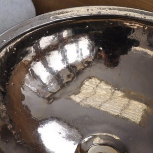 K&ouml;nnte beinhalten: Nahaufnahme einer geh&auml;mmerten Metallschale mit einer reflektierenden, rissigen Oberfl&auml;che. Die Schale hat einen breiten Rand und ein dunkles, strukturiertes Inneres. Das Metall hat eine silberne Farbe und reflektiert das Licht.