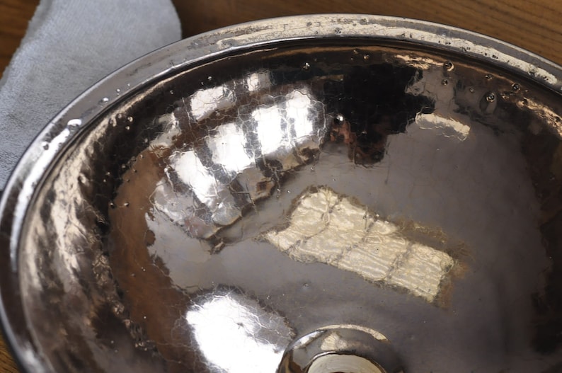 K&ouml;nnte beinhalten: Nahaufnahme einer geh&auml;mmerten Metallschale mit einer reflektierenden, rissigen Oberfl&auml;che. Die Schale hat einen breiten Rand und ein dunkles, strukturiertes Inneres. Das Metall hat eine silberne Farbe und reflektiert das Licht.