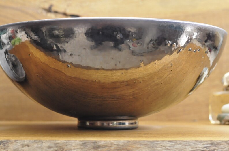 K&ouml;nnte beinhalten: Handgefertigte Keramikschale mit metallisch silberner und goldener Oberfl&auml;che. Die Schale hat eine breite, flache Form und steht auf einem kleinen, kreisf&ouml;rmigen Sockel. Das Innere reflektiert das Licht und erzeugt einen einzigartigen visuellen Effekt.