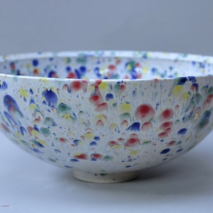 May include: A white ceramic bowl with a speckled design. The bowl is decorated with colorful paint splatters in red, blue, yellow, and green. The bowl has a rounded shape and sits on a small, circular base.