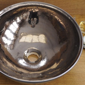 K&ouml;nnte beinhalten: Ein Waschbecken aus geh&auml;mmertem Metall mit reflektierender Oberfl&auml;che. Das Waschbecken ist rund mit einem Abflussloch in der Mitte. Ein St&uuml;ck Seife liegt auf einer dekorativen Seifenschale, und eine Parf&uuml;mflasche steht daneben.
