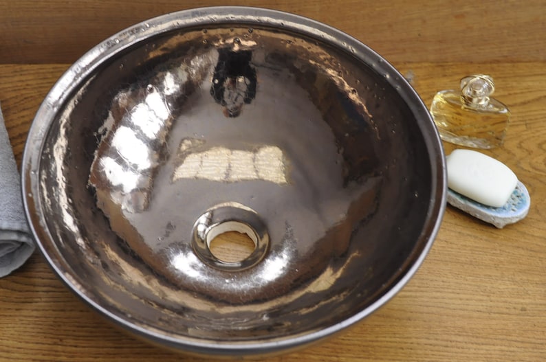 K&ouml;nnte beinhalten: Ein Waschbecken aus geh&auml;mmertem Metall mit reflektierender Oberfl&auml;che. Das Waschbecken ist rund mit einem Abflussloch in der Mitte. Ein St&uuml;ck Seife liegt auf einer dekorativen Seifenschale, und eine Parf&uuml;mflasche steht daneben.