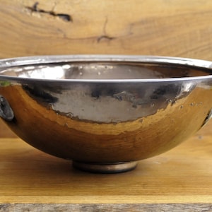 K&ouml;nnte beinhalten: Eine metallisch silberfarbene Sch&uuml;ssel mit runder Form und kleinem, kreisf&ouml;rmigem Sockel. Die Sch&uuml;ssel hat eine reflektierende Oberfl&auml;che und steht auf einer Holzoberfl&auml;che. Ein zusammengerolltes graues Handtuch, ein St&uuml;ck Seife und eine Parf&uuml;mflasche befinden sich im Hintergrund.