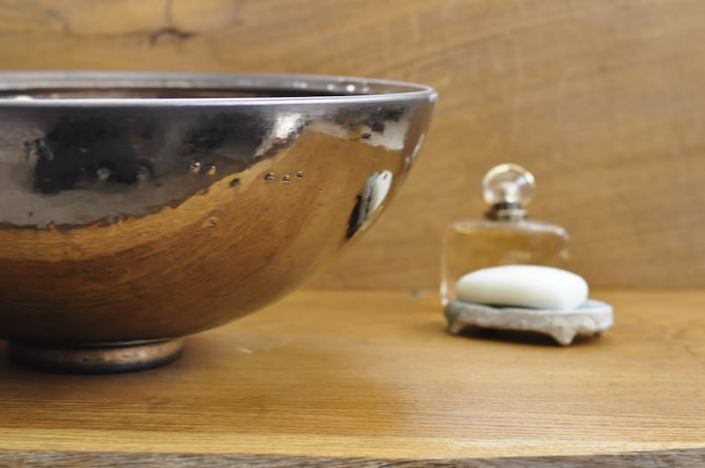 K&ouml;nnte beinhalten: Eine metallisch braune Keramikschale mit dunklem Rand steht auf einer Holzoberfl&auml;che. Im Hintergrund befinden sich eine kleine Seifenschale mit einem Seifenst&uuml;ck und eine Parf&uuml;mflasche. Rustikale Badezimmerdekoration.