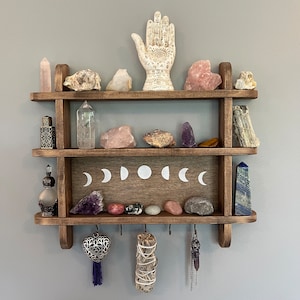 May include: Two-tiered wooden shelf with a white ceramic hand, crystals, and other decorative items. The shelf has a moon phase design and hooks for hanging items.