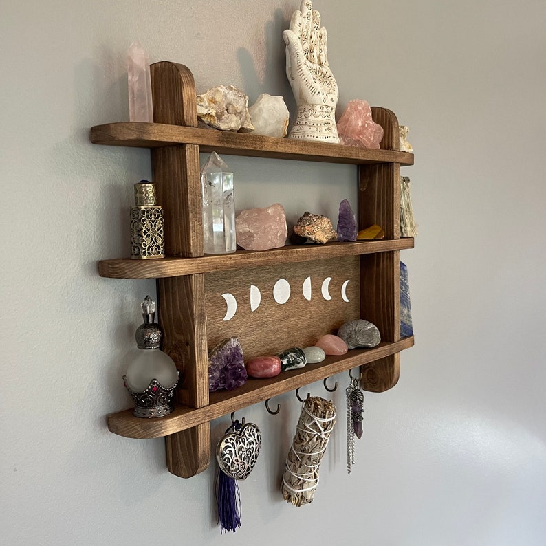 May include: Wooden shelf with three tiers, featuring a hand-painted moon phase design. The shelf is decorated with crystals, a hand sculpture, and other decorative items.