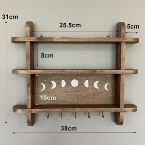 May include: A dark brown wooden shelf with two shelves and five hooks on the bottom. The top shelf is 25.5 inches wide and the bottom shelf is 10 inches wide. The shelf is 31 inches tall and 38 inches wide. The shelf has a moon phase design on the front.