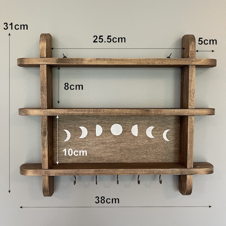 May include: A dark brown wooden shelf with two shelves and five hooks on the bottom. The top shelf is 25.5 inches wide and the bottom shelf is 10 inches wide. The shelf is 31 inches tall and 38 inches wide. The shelf has a moon phase design on the front.