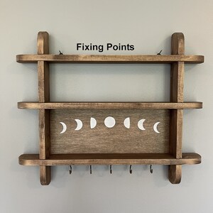 May include: A wooden shelf with two shelves and hooks on the bottom. The shelf has a moon phase design and the text "Fixing Points" above the top shelf.