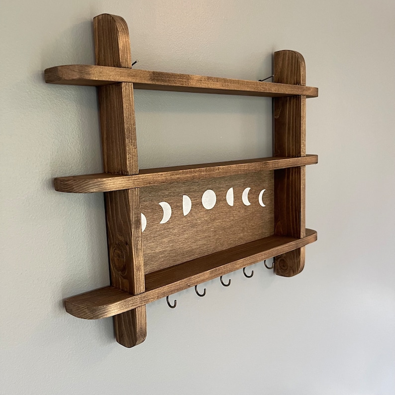 May include: A wooden shelf with two shelves and a row of hooks underneath. The shelf has a moon phase design painted on the front.