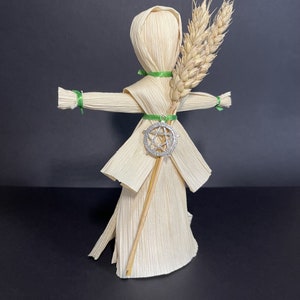 May include: A handmade corn husk doll with a green ribbon around its waist and a silver pendant. The doll is holding a bundle of wheat stalks.
