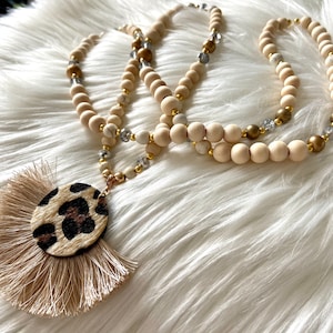 Peut inclure: Un long collier à plusieurs rangs avec des perles en bois, des perles dorées et des perles de cristal transparentes. Le collier a un grand pendentif avec un cercle imprimé léopard brun et beige et un pompon beige.