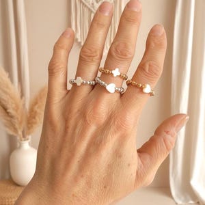 May include: A hand displays several beaded rings. The rings feature heart, cross, and flower designs, with silver and gold beads. The rings are on a hand with a neutral background.