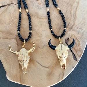 Pode incluir: Dois colares de ouro com contas pretas e um pingente de crânio de touro dourado. O pingente de crânio de touro tem chifres pretos.