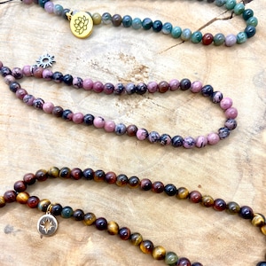 May include: Three beaded necklaces with different colored stones and charms. The top necklace has green and brown stones with a gold flower charm. The middle necklace has pink and black stones with a silver sun charm. The bottom necklace has brown and gold stones with a silver star charm.