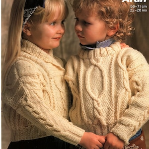 May include: Two children wearing cream coloured cable knit sweaters. The girl is wearing a blue bandana in her hair. The boy is wearing a brown bandana around his waist. The pattern is called "Aran" and the size is 56-71 cm or 22-28 inches.