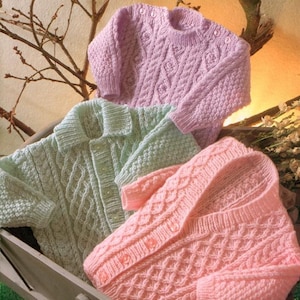 May include: Three knitted baby sweaters in pink, green and light purple. The sweaters have a cable knit pattern and button fastenings. The sweaters are displayed in a wooden crate.