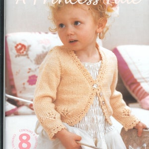 May include: A young girl wearing a peach-colored knitted cardigan with a white button closure. The cardigan has a delicate lace trim. The girl is holding a silver wand and wearing a white dress. The image includes the text "A Princess Tale" and "Fairytale 4ply & DK 16-26in (41-66cm)".