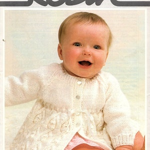May include: A white knitted baby cardigan with a button fastening and a tie at the waist. The cardigan is made with a delicate stitch pattern and has a scalloped edge. The baby is wearing a pink and white gingham nappy cover. The pattern number is 13432, and the size is 16-20 inches or 41-51 cm.