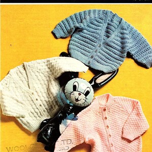 May include: Three knitted baby cardigans in blue, white, and pink. The cardigans are laid out on a yellow background with a toy bunny. The pattern is Copley Lotus Baby Golden Sovereign 4 ply. The pattern number is 1124 and the price is 10p. The size is 18-20 inches or 46-51 cm.