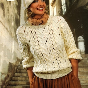 May include: A white knitted sweater with a cable knit pattern and a lace stitch design. The sweater has a round neckline and long sleeves. The model is wearing a white beret and a brown scarf.