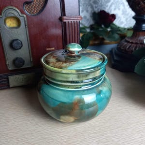 May include: A ceramic jar with a lid, featuring a blue, green, and brown glaze. The jar has a rounded shape and a smooth, glossy finish.
