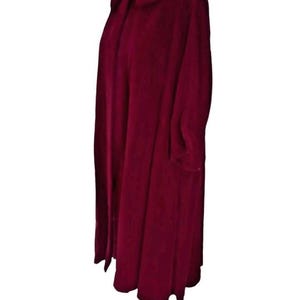May include: A long, burgundy-colored coat with a wide collar and long sleeves. The coat is displayed on a wooden dress form. The fabric appears to be a soft, possibly velvet, material. The coat has a flowing, full shape.