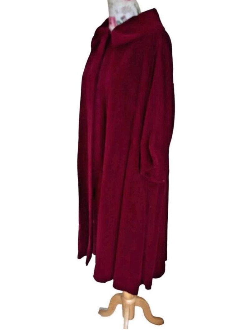 May include: A long, burgundy-colored coat with a wide collar and long sleeves. The coat is displayed on a wooden dress form. The fabric appears to be a soft, possibly velvet, material. The coat has a flowing, full shape.
