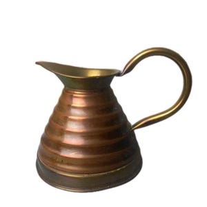 May include: A small, vintage copper jug with a brass handle and spout. The body of the jug has a ribbed design, and the handle curves upwards. The jug is likely used for serving liquids and has a classic, antique aesthetic.