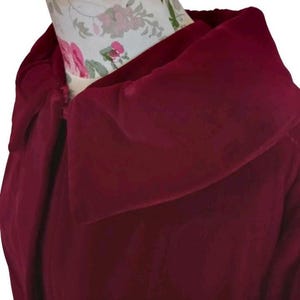May include: A close-up of a burgundy-colored garment with a wide, draped collar. The fabric appears to be soft and flowing, with a slight sheen. The collar is the focal point, creating a dramatic effect. The garment is displayed on a mannequin.