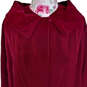 May include: A deep red velvet coat with a large collar. The coat is buttoned at the neck and appears to be vintage. The fabric has a soft, luxurious texture. The coat is displayed on a mannequin with a floral pattern.