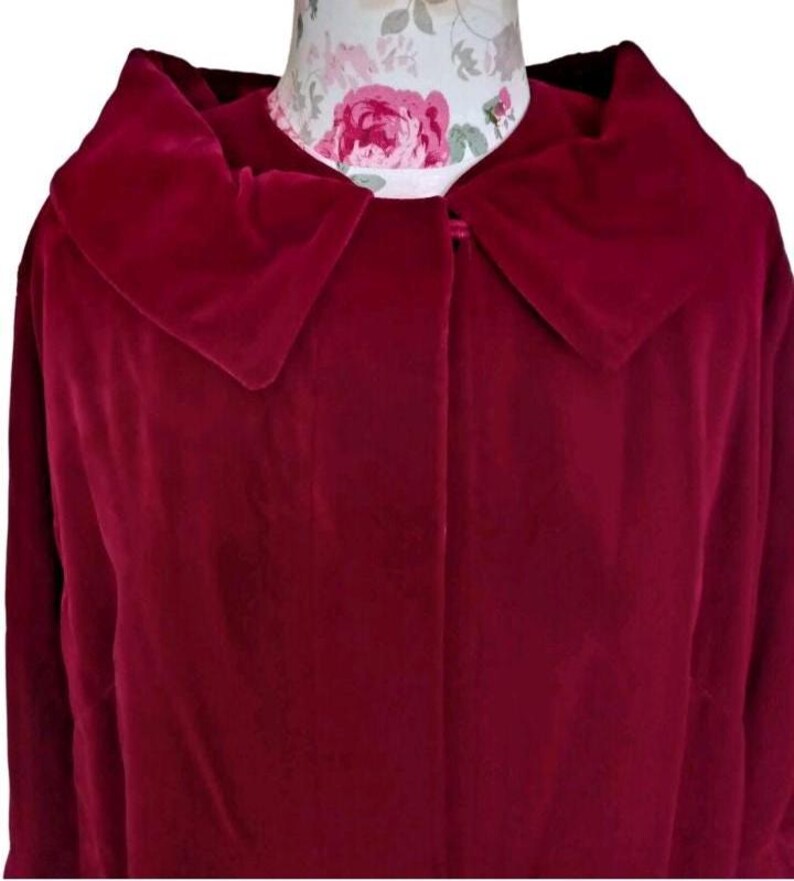 May include: A deep red velvet coat with a large collar. The coat is buttoned at the neck and appears to be vintage. The fabric has a soft, luxurious texture. The coat is displayed on a mannequin with a floral pattern.