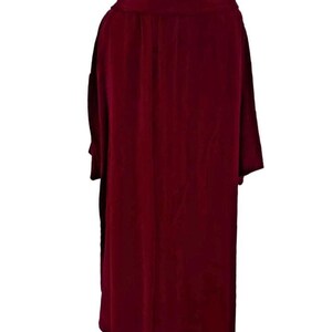 May include: A long, burgundy-colored velvet coat with a wide collar and long sleeves. The coat is displayed on a wooden hanger, showcasing its flowing design and rich color. The fabric appears soft and drapes elegantly.