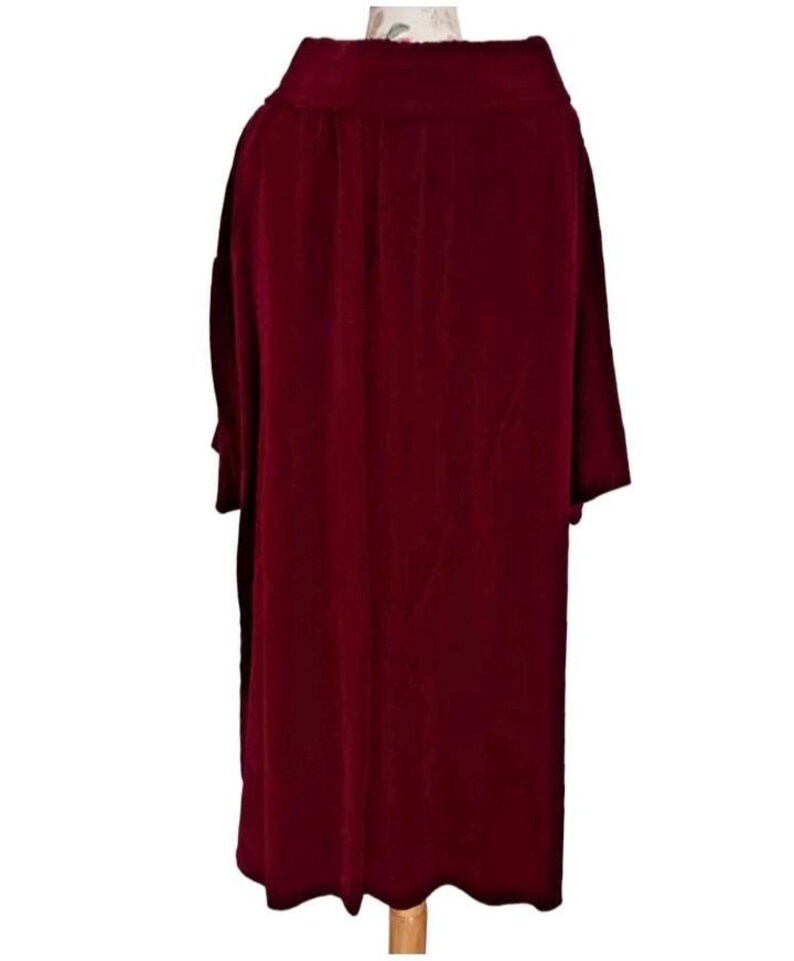 May include: A long, burgundy-colored velvet coat with a wide collar and long sleeves. The coat is displayed on a wooden hanger, showcasing its flowing design and rich color. The fabric appears soft and drapes elegantly.