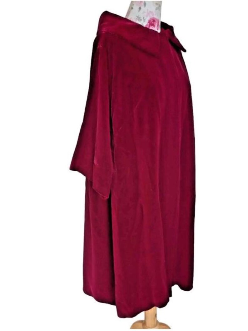 May include: A deep burgundy velvet cape with a wide collar and short sleeves. The cape is draped over a wooden mannequin, showcasing its flowing design. The fabric has a soft, luxurious texture, and the color is rich and vibrant.