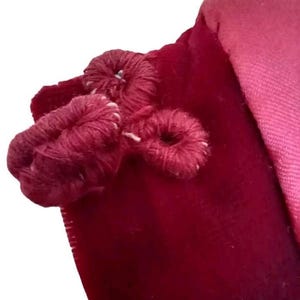 May include: Close-up of a deep red velvet fabric with three decorative, looped yarn embellishments. The yarn is a darker shade of red, and the stitching is visible. A lighter pink fabric is visible in the background.