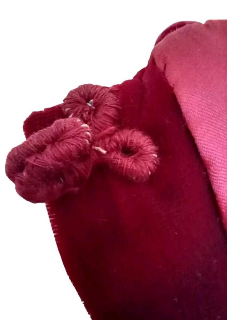 May include: Close-up of a deep red velvet fabric with three decorative, looped yarn embellishments. The yarn is a darker shade of red, and the stitching is visible. A lighter pink fabric is visible in the background.
