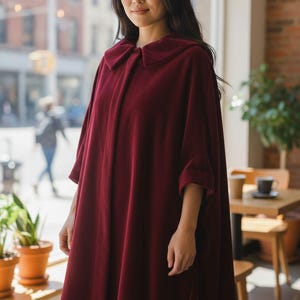 May include: A burgundy velvet cape with a rounded collar and three-quarter sleeves. The cape is knee-length and worn by a person in a cafe setting, with natural light from a large window.