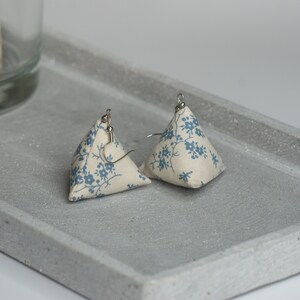 Puede incluir: Un par de pendientes triangulares de tela con un patrón floral azul y blanco. Los pendientes cuelgan de ganchos plateados.