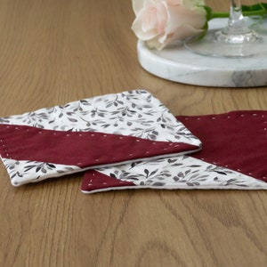 Könnte beinhalten: Zwei Untersetzer aus Stoff in Burgunderrot und Weiß mit einem Blattmotiv. Die Untersetzer haben einen diagonalen burgunderfarbenen Streifen mit weißen Nähten.