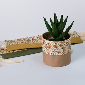 Peut inclure: Une plante succulente verte dans un pot en tissu floral blanc et brun avec une base en tissu brun. Le pot a deux sangles en tissu avec des liens blancs.