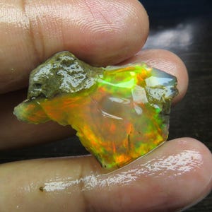 Puede incluir: Una piedra preciosa de ópalo en bruto sin pulir que presenta un cuerpo translúcido con destellos vibrantes de verde neón, naranja y amarillo. La piedra tiene una forma irregular y dentada con una matriz rugosa de color gris parduzco.
