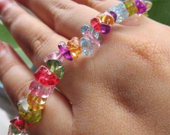 Op de afbeelding: Een elastische armband bestaande uit talloze kleine, onregelmatige, getrommelde edelsteensplinters in een levendig kleurenpalet, waaronder doorschijnend roze, felrood, limoengroen, zonnig geel, oranje en lichtblauw, gerangschikt in een doorlopende streng.