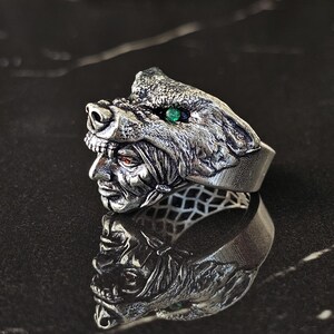 May include: A silver-toned ring featuring a detailed wolf head design with a green gemstone eye. The ring also depicts a human face with red eyes. The ring is displayed on a reflective surface.