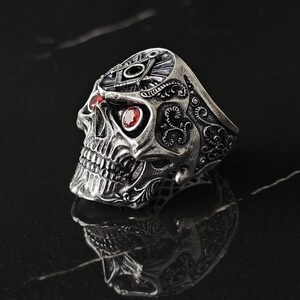 May include: A silver skull ring with red gemstone eyes. The ring has detailed engravings and a design on the forehead. The skull's jaw is visible, and the ring is set against a dark background.