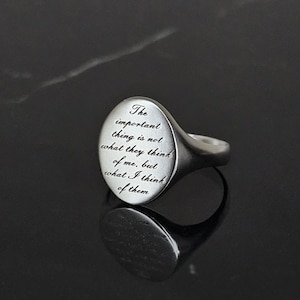 Może przedstawiać: Srebrny pierścień sygnetowy z owalnym wierzchołkiem, na którym wygrawerowano cytat: "Ważne jest nie to, co oni myślą o mnie, ale to, co ja myślę o nich."