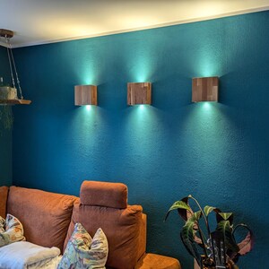 Könnte beinhalten: Ein Wohnzimmer mit einer türkisblauen Wand, an der drei quadratische Holz-Wandleuchten angebracht sind. Darunter befindet sich ein orangefarbenes Ecksofa mit gemusterten Kissen. Ein hängendes Holzregal mit einer Pflanze und eine Bodenpflanze ergänzen das Bild.
