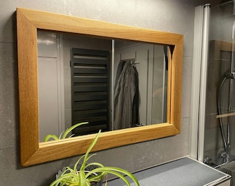 Peut inclure: Un miroir rectangulaire avec un cadre en bois. Le miroir est accroché à un mur en carrelage gris. La réflexion dans le miroir montre une salle de bain avec un porte-serviettes et une douche.