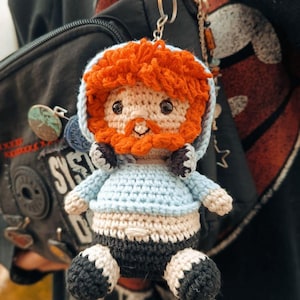 Puede incluir: Un llavero de ganchillo hecho a mano con una pequeña muñeca con pelo y barba naranja. La muñeca lleva una chaqueta con capucha azul claro y pantalones cortos negros. El llavero está sujeto a un mosquetón rojo. La muñeca tiene un tono de piel neutro.